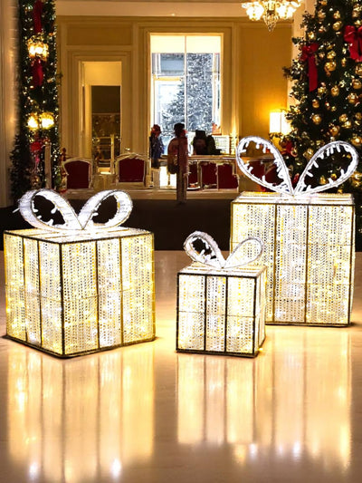 Set of three indoor LED gift boxes glowing in warm white with pure white ribbon accents, displayed in a grand holiday lobby with Christmas trees and garland