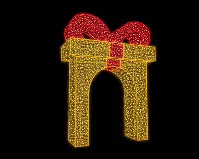 Walk-through gift box arch lit with warm white LED lights and topped with a red ribbon, installed outdoors as part of a nighttime holiday light display