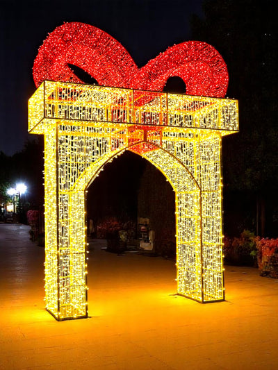 Walk-through gift box arch lit with warm white LED lights and topped with a red ribbon, installed outdoors as part of a nighttime holiday light display