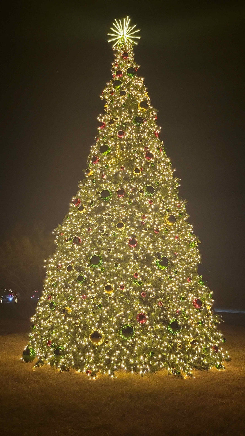 Illuminated commercial Christmas tree at night with warm white LED lights and oversized red, green, and gold ornaments
