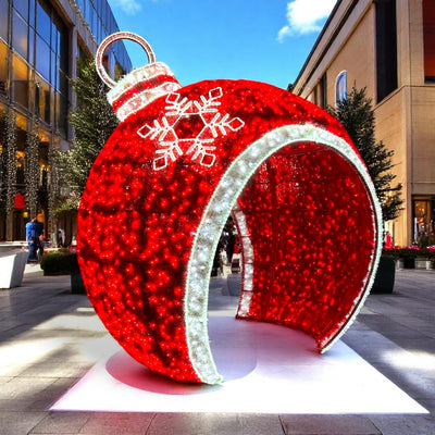 Oversized walk-through red LED ornament sculpture with white snowflake accents and glowing archway, featured in a commercial outdoor holiday display